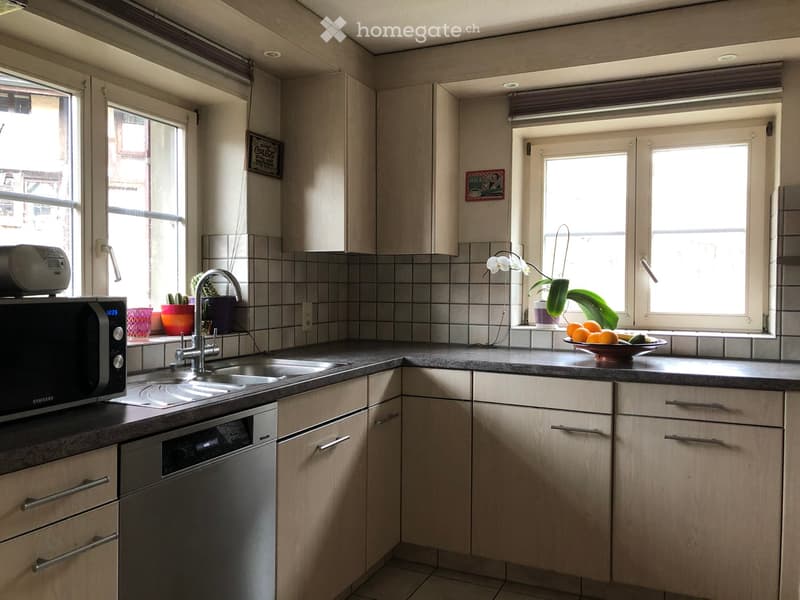 Modern kitchen with light wood cabinets, stainless steel appliances, and a black countertop. Features a double sink, microwave, and decorative items on the windowsill.