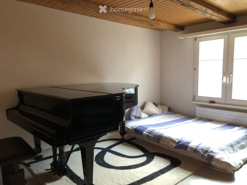 Cozy bedroom with a double bed, piano, and large windows letting in natural light. The room features a patterned rug and exposed wooden beams on the ceiling.