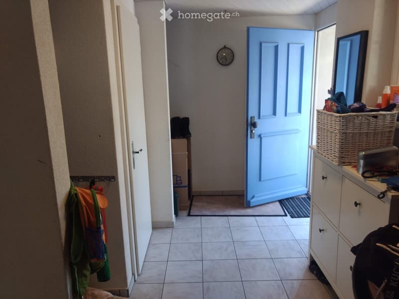 Hallway with tiled floor, white walls, a blue door at the end, a coat rack with jackets, a small table with shoes, a clock on the wall, and a wicker basket on a cabinet.