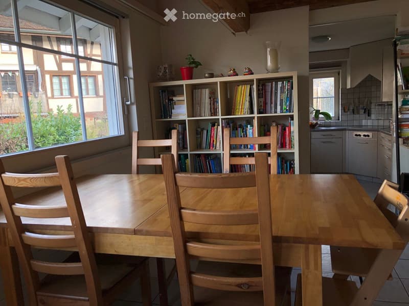Bright room with large windows, wooden dining table with chairs, bookshelf filled with books, and a small kitchen area with a dishwasher.