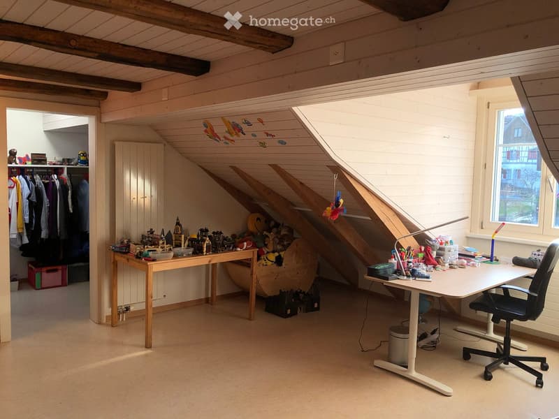 Bright, slanted-ceiling room with two desks, one with a globe and lamp, the other with toys and books, a cabinet, and a closet with clothes.