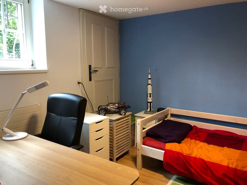 A small, well-lit bedroom with a single bed, a desk with a chair, a lamp, and a toy car on a small table. The walls are painted in two colors, and a rocket model is displayed on a side table.