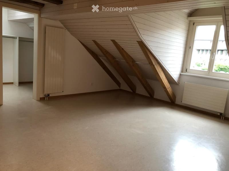 Empty attic room with wooden slanted ceilings, white walls, large window, and radiator.