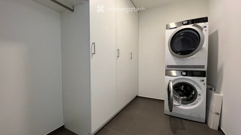 A small, modern laundry room with a stacked washer and dryer unit, white cabinetry, and dark tiled flooring.