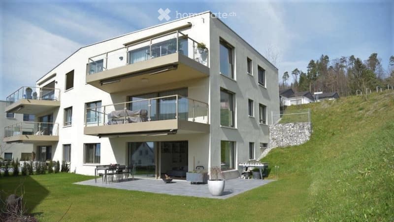 Modern two-story house with large windows, multiple balconies, and a raised ground floor. Features a spacious outdoor patio with a dining set, grill, and potted plants.