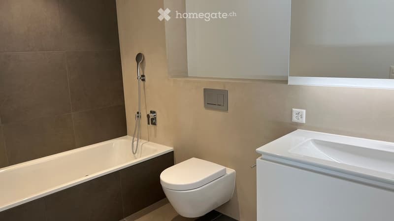 Modern bathroom with a bathtub, toilet, sink, and showerhead. Beige tiles and a large mirror are visible.