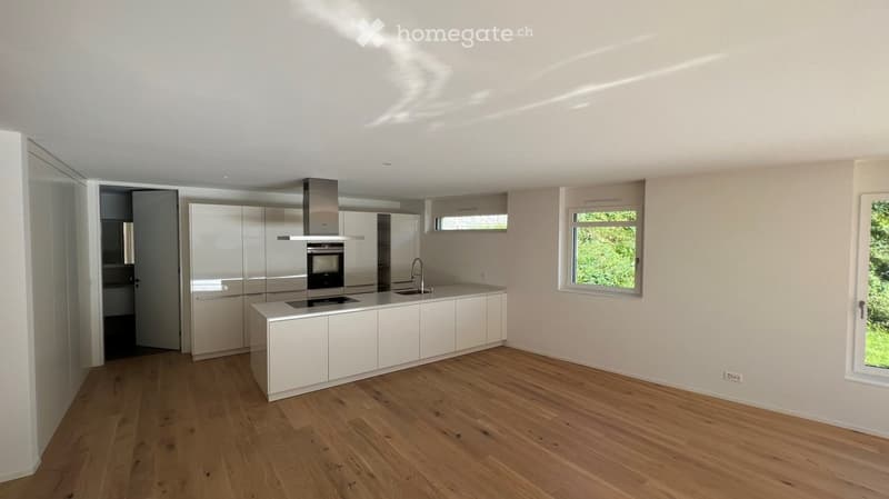 Modern, minimalist kitchen with white cabinetry, a central island, and a built-in oven and range hood. The space features light wooden flooring and large windows allowing natural light to fill the room.