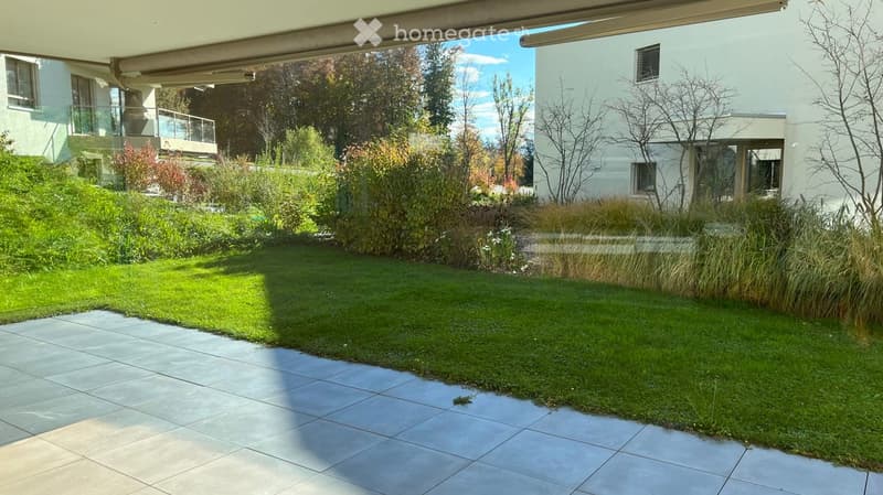 Well-maintained garden with a tiled patio, green lawn, and various plants and shrubs, with modern buildings in the background.