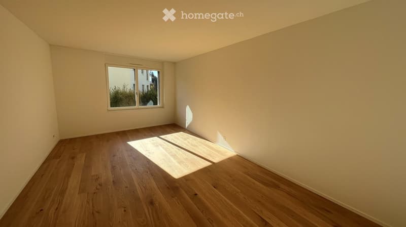 Empty room with light wooden flooring and white walls, featuring a single window with a view of greenery outside.
