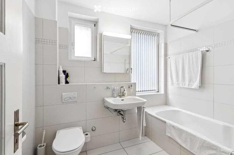 Modern bathroom with white tiles, a sink, toilet, bathtub, mirror, and towel. Features a window with blinds and a small cabinet with toiletries.