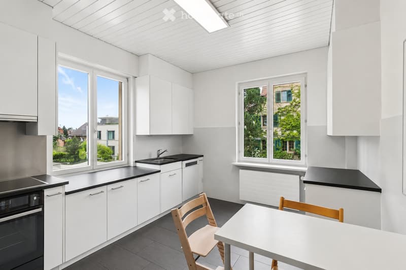 Modern kitchen with white cabinets, black countertops, and large windows overlooking a green area. Includes a sink, oven, and a small dining area with a table and chairs.