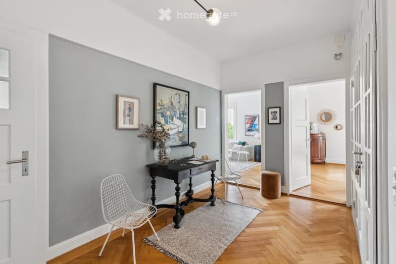 Modern hallway with wooden flooring, grey walls, a small black table with a vase and picture frames, white wire chairs, and an open doorway leading to a living area with a sofa and additional decor.