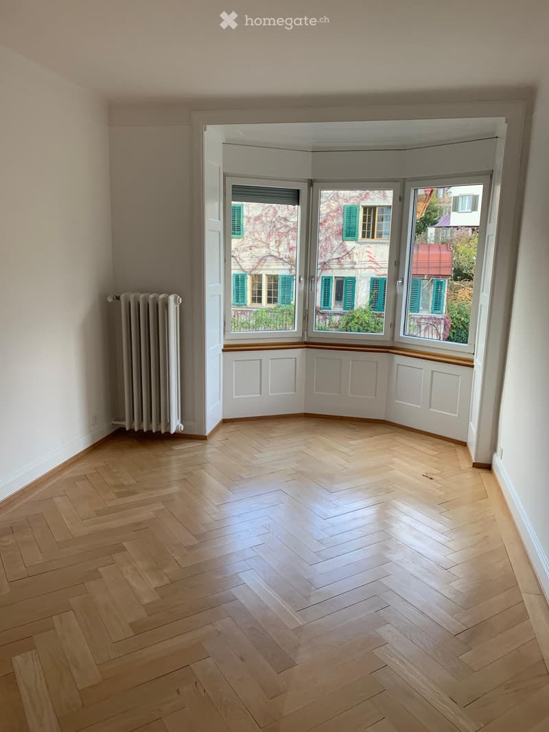 Empty room with wooden herringbone flooring, white walls, and large windows with shutters opening to a view of neighboring buildings.