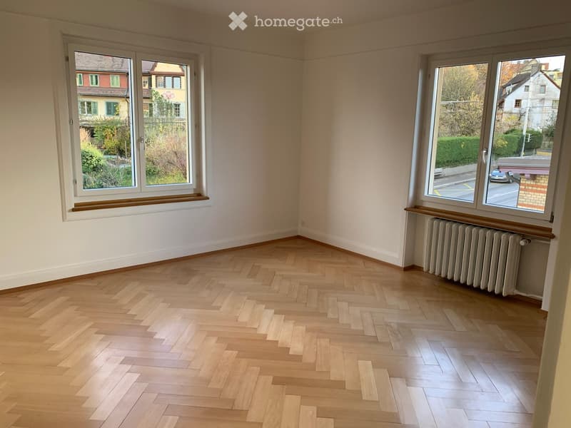 Empty room with wooden herringbone flooring, white walls, and large windows offering views of neighboring houses and a street.
