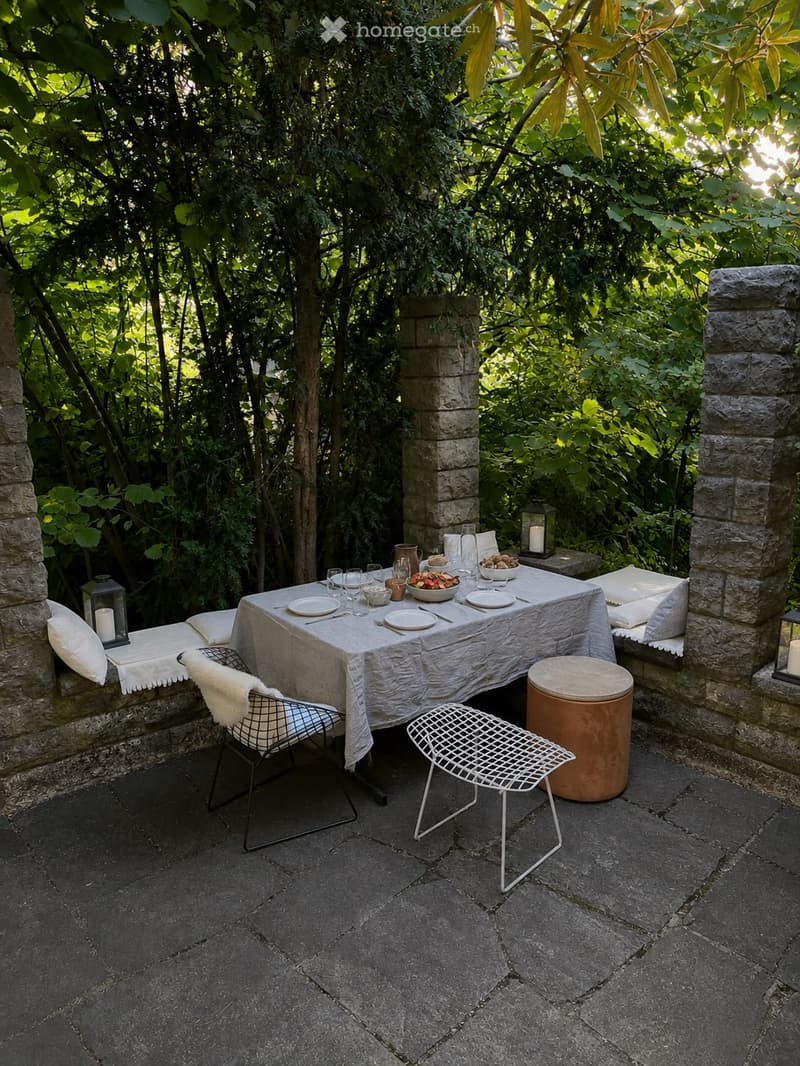 An outdoor dining area surrounded by lush greenery, featuring a stone-paved floor, a long dining table with plates, bowls, and glasses, two modern wire chairs, a round stool, and lanterns on the stone walls.