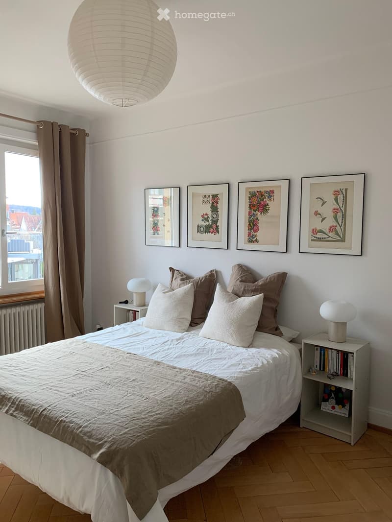 Cozy bedroom with a large bed, multiple pillows, a nightstand with a lamp, a mirror, and several framed pictures on the wall.