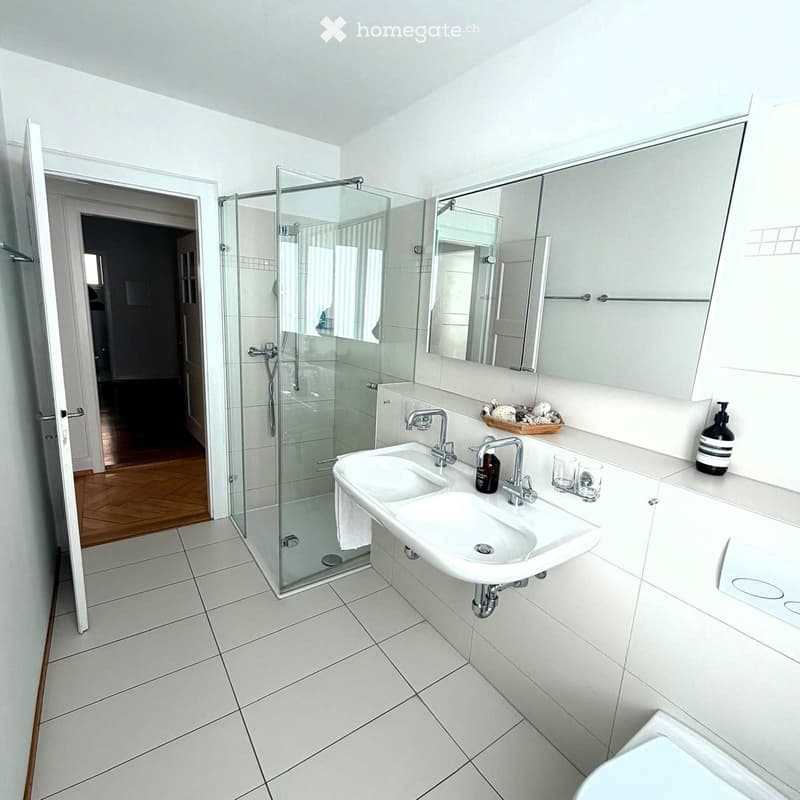 Modern bathroom with white tiles, a double sink, a glass-enclosed shower, and a large mirror above the sink.