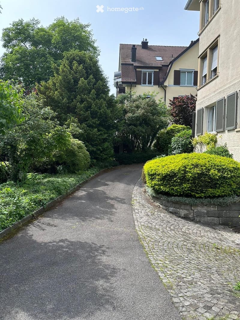 A driveway leads to a two-story house with a red-tiled roof and multiple windows. The surrounding area is landscaped with trees, shrubs, and a well-maintained garden.