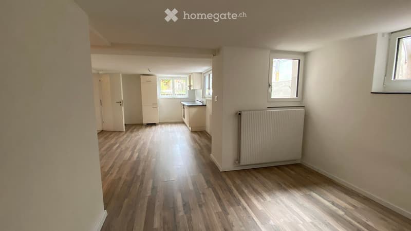 Empty room with wooden flooring, white walls, and a radiator. The room is connected to a small kitchen area with cabinets and a sink.