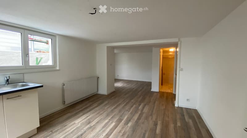 Empty room with wooden flooring, white walls, and a small sink area. A doorway leads to another room with warm lighting.