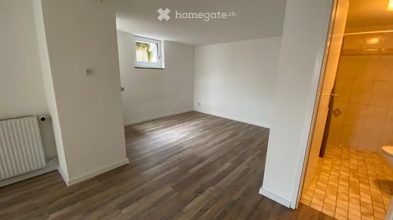 Empty room with wooden flooring, white walls, a radiator, and a window. Adjacent bathroom with tiled walls and a toilet is visible.