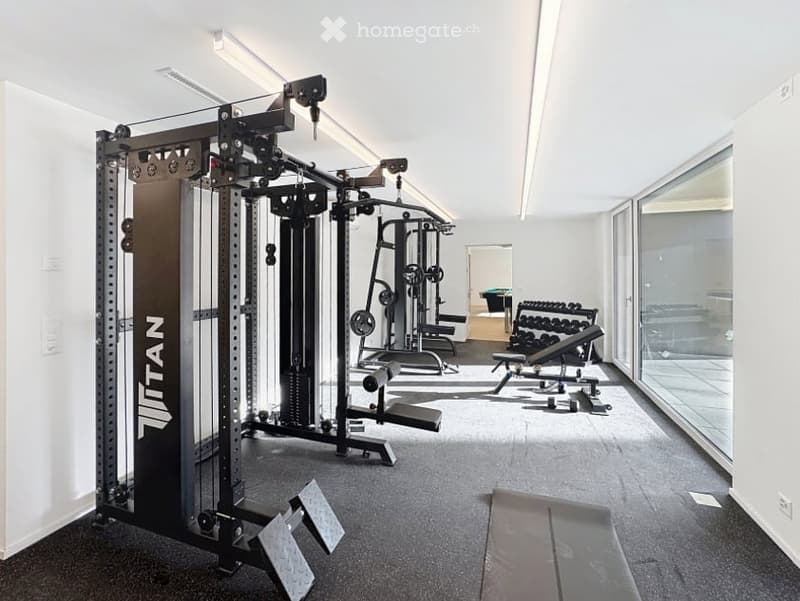 Modern gym room with a large multi-gym machine, bench press, dumbbells, weight plates, and a treadmill. The space is well-lit with large windows and a clean, organized layout.