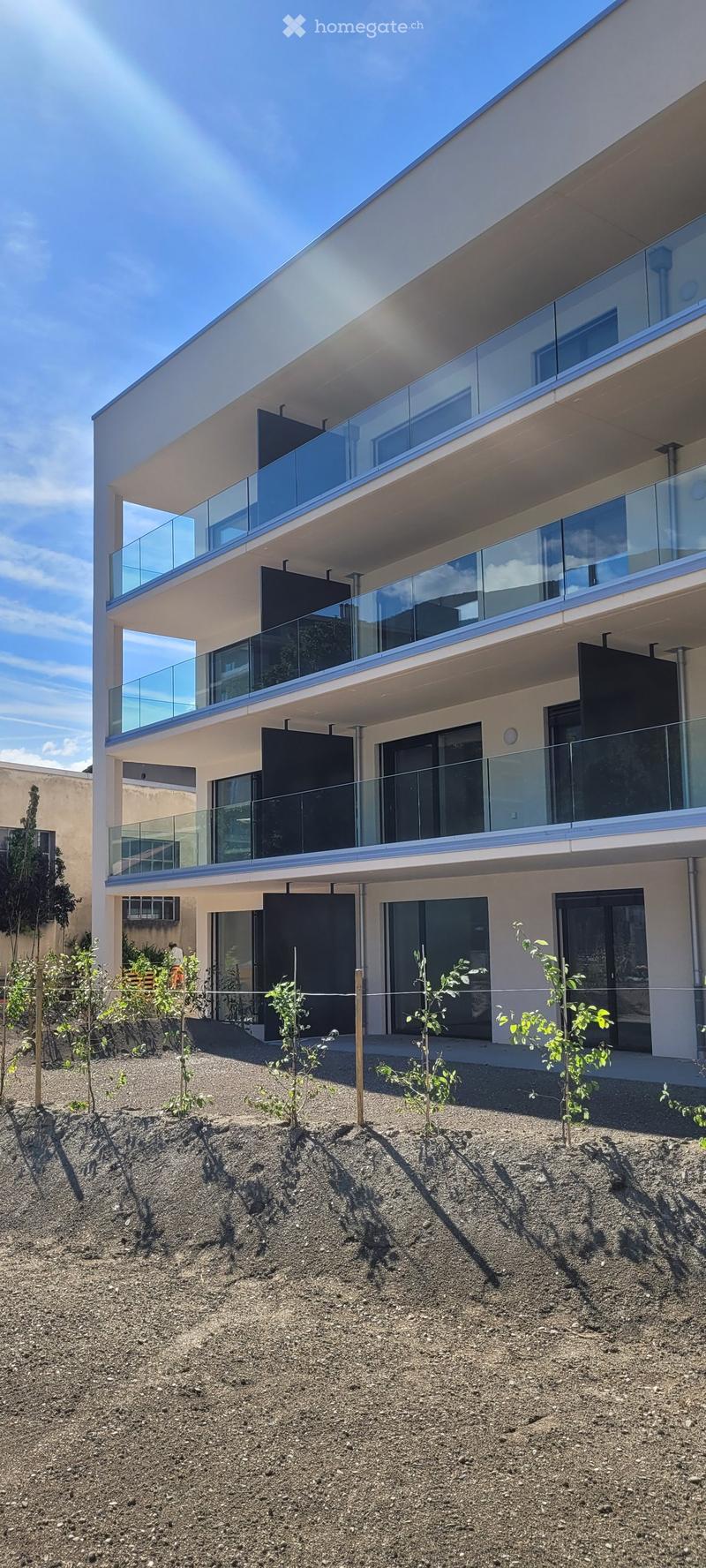 Modern multi-story building with large glass balconies and contemporary design, surrounded by small plants and gravel ground.