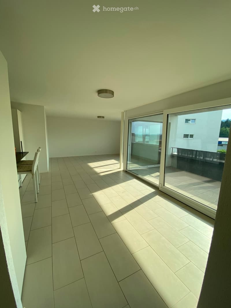 Bright, unfurnished room with tiled floor, large sliding glass doors leading to a balcony, and minimal furniture including a single chair and small table.