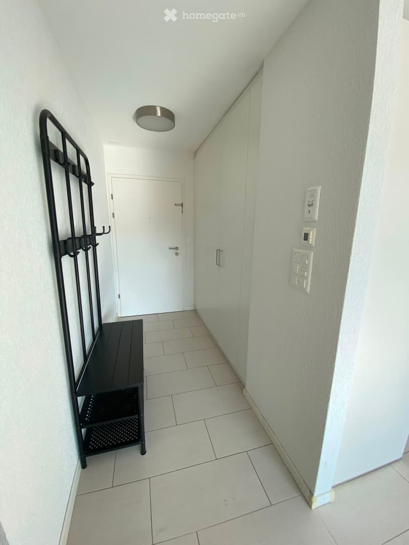 A clean, modern hallway with white tiled flooring, light grey walls, and a white door at the end. A black bench and coat rack are positioned against the left wall, and a round ceiling light fixture is mounted above.