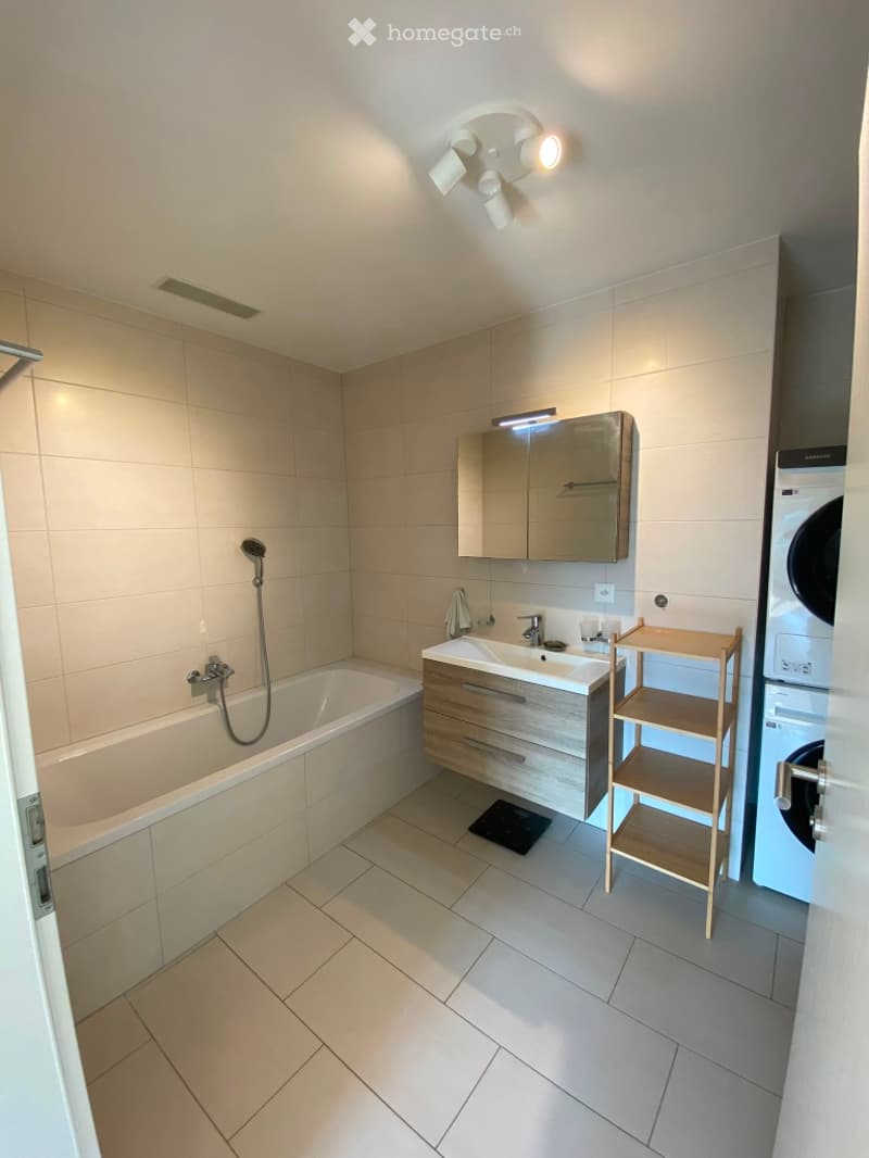 Modern bathroom with a bathtub, sink, mirror cabinet, wooden shelf, and a private washing machine. Light-colored tiles and a sleek design.