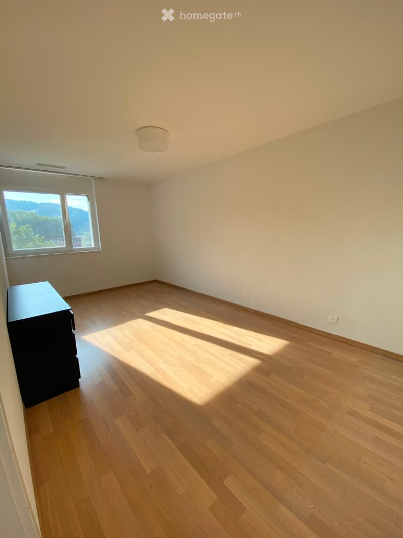 Empty room with wooden flooring, white walls, and a large window letting in natural light. A small black cabinet is visible against one wall.
