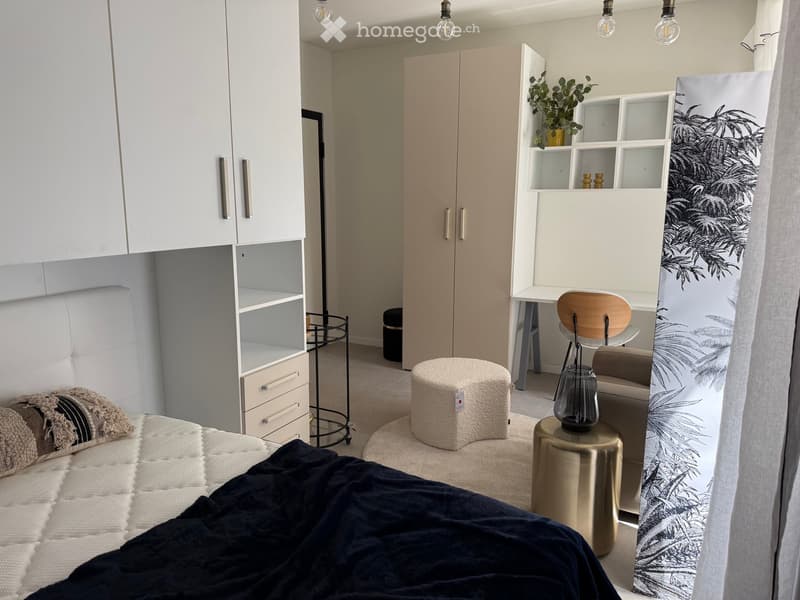 Modern bedroom with a white bed, dark blue blanket, white cabinets, a round stool, a wooden chair, a small table with a lamp, and a decorative shelf with plants.