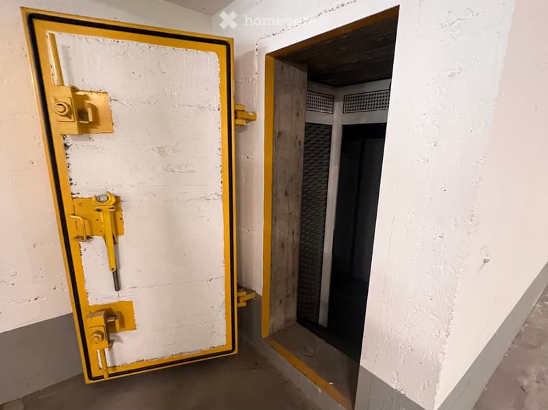 A narrow corridor with white walls and a concrete floor, featuring a large yellow metal door on the left and a small open doorway on the right leading to another room.