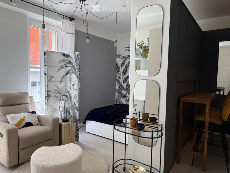 Modern, minimalist room with a sofa, bed, small table, and chairs. Features large mirrors, pendant lights, and a decorative plant.