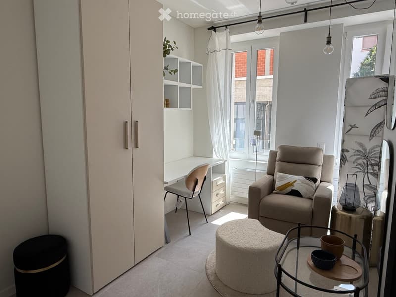 A modern, well-lit living space with a sofa, round stool, glass coffee table, chair, and a small desk area with a chair and shelves.