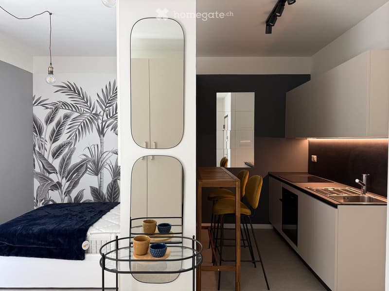 Modern studio apartment with a bed, small table, chairs, and a compact kitchen area with a sink and cabinets.