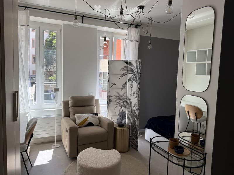 Modern living room with a beige sofa, round white stool, glass table, and decorative items. Large windows with white curtains and a view of greenery outside.