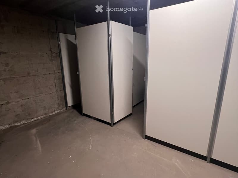 Empty restroom area with concrete walls, a concrete floor, and three stall partitions with metal frames and white panels.