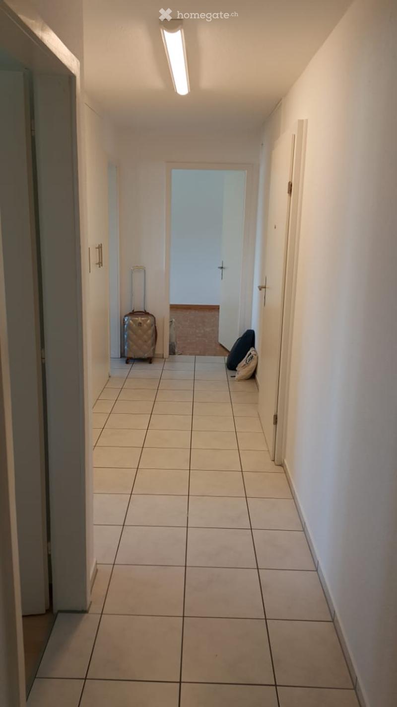 A narrow hallway with tiled flooring, white walls, and a single overhead light. A suitcase and bags are placed near the end of the hallway.