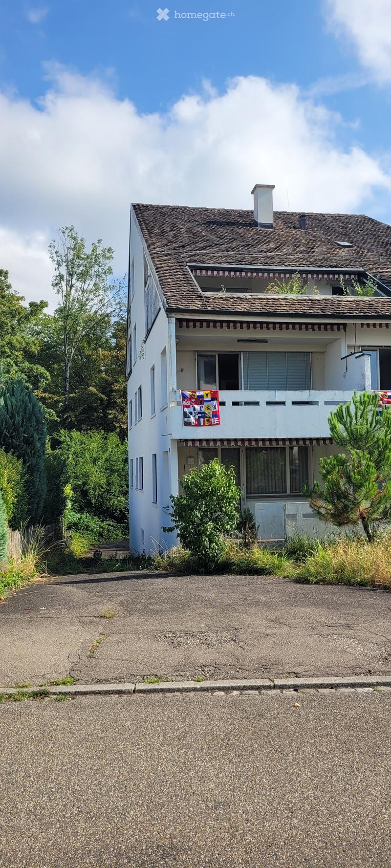 Two-story house with a tiled roof, white exterior, and a balcony adorned with a colorful flag. The house is surrounded by greenery and has a paved driveway.