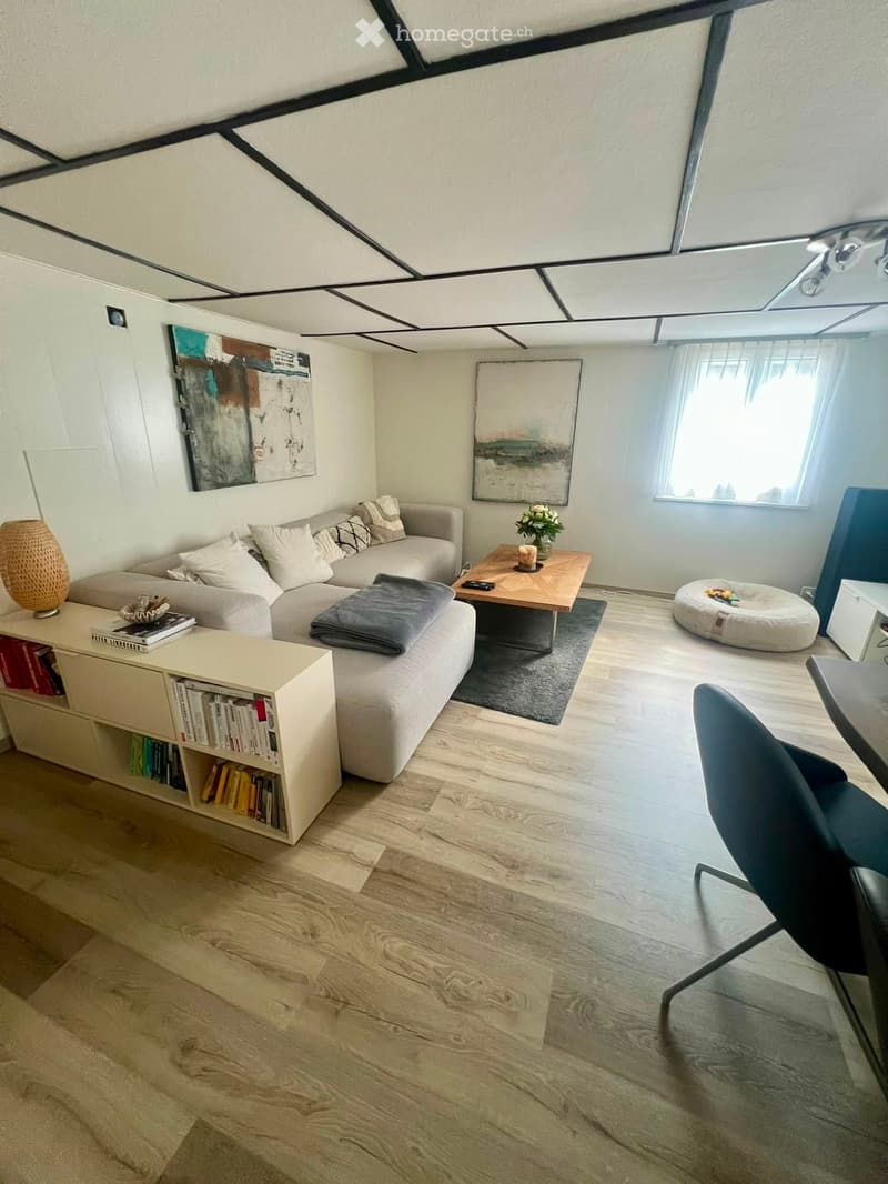 Modern living room with light-colored wooden flooring, a grey sectional sofa, a wooden coffee table, a bookshelf, and a round ottoman.