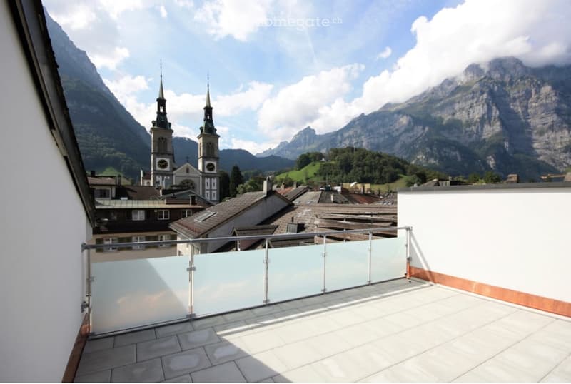 Balcony with glass railing, tiled floor, overlooking a town with church towers and mountain backdrop.