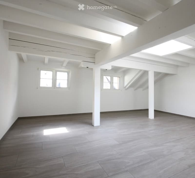 Completely empty room with white walls, wooden floor, and exposed white beams. No furniture or equipment.