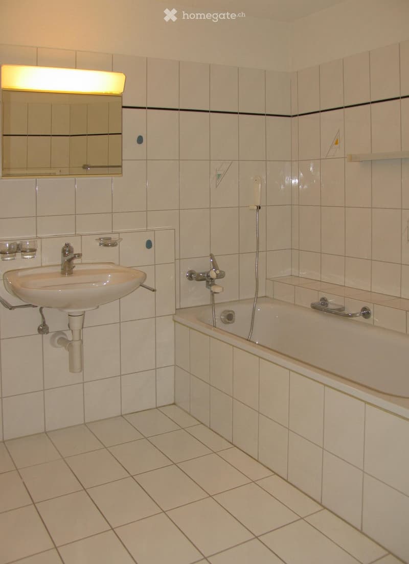 Bathroom with white tiled walls and floor, featuring a sink with a mirror above it, a bathtub with a handheld showerhead, and a built-in shelf on the wall.