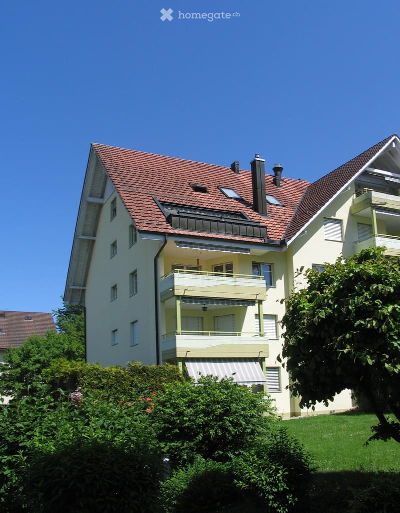 Two-story house with red-tiled roof, multiple windows, balconies, and surrounding greenery.
