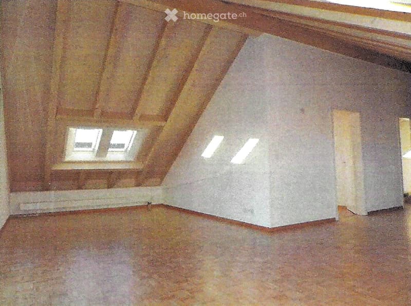 Empty attic room with wooden slanted ceilings, exposed beams, and skylights. Bare wooden floor and white walls.