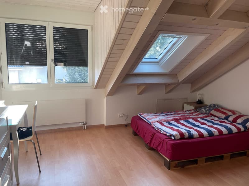 Cozy attic bedroom with wooden slanted ceilings, skylight windows, a bed with a red and blue striped blanket, a small table with a chair, and a nightstand with a lamp.