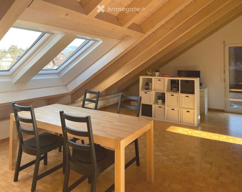 Bright attic room with wooden beams, skylights, a wooden table with four chairs, a white shelving unit with baskets and small plants, and a wooden floor.