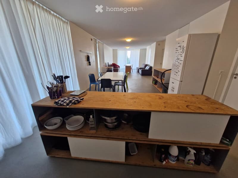 Open-plan living and dining area with a kitchen island, sofa, dining table, and chairs. The kitchen features a wooden island with bowls, plates, and cleaning supplies on the shelves.