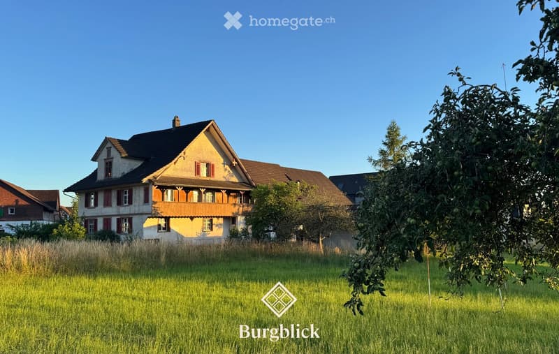 A two-story house with a pitched roof, wooden balcony, and red shutters, surrounded by a grassy field and trees.